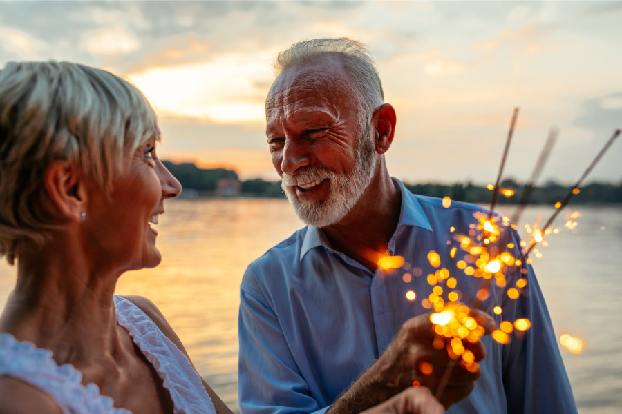 A white senior couple smiles as they hold sparklers outside by the sea together